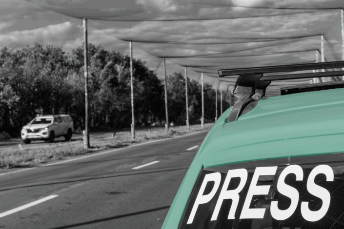 A black and white image of a road with the back of an SUV in the foreground. The SUV has the words 'PRESS' emblazoned on the back windshield.